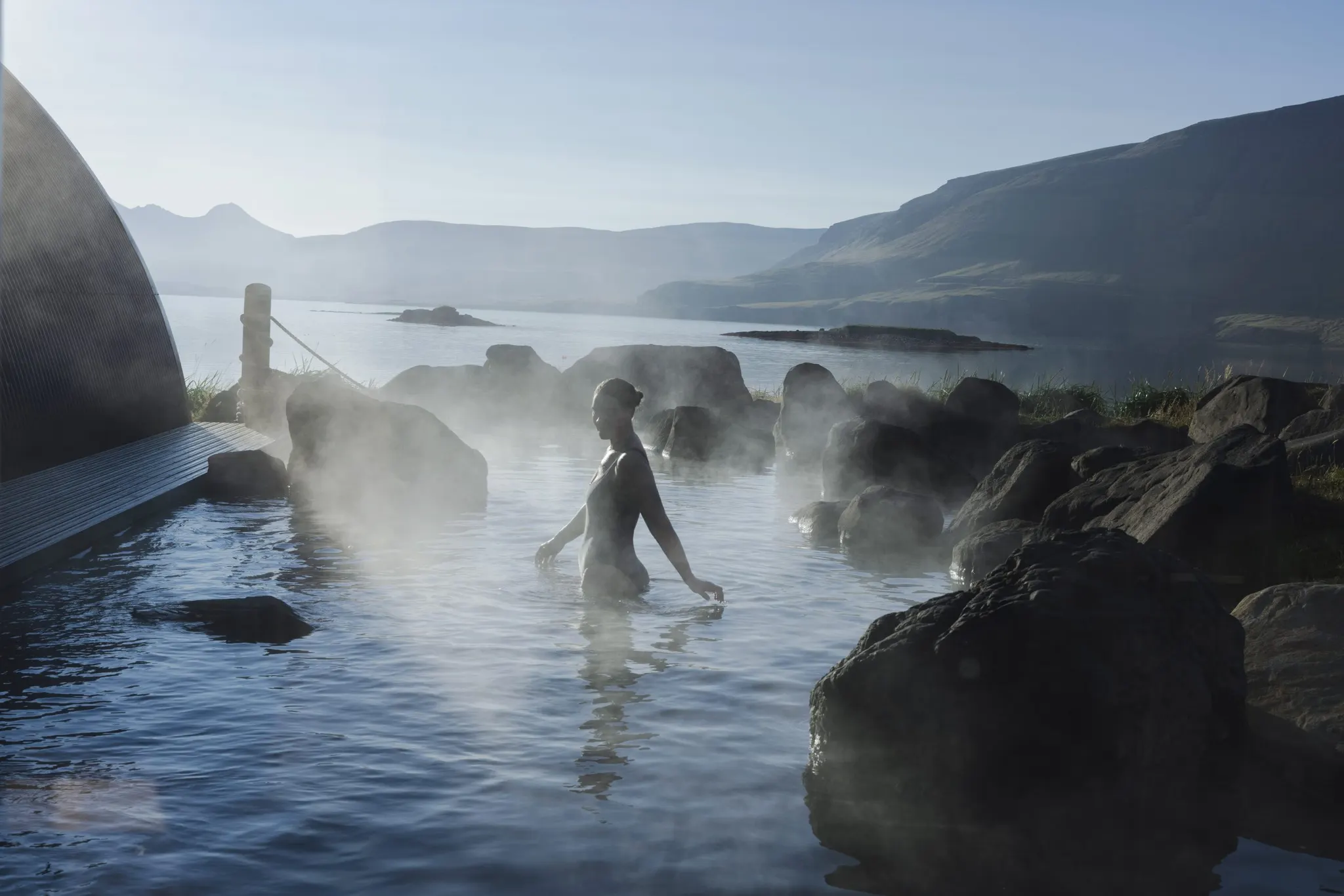 A steaming pool with a lone bather and the mountains of the Whale Fjord behind