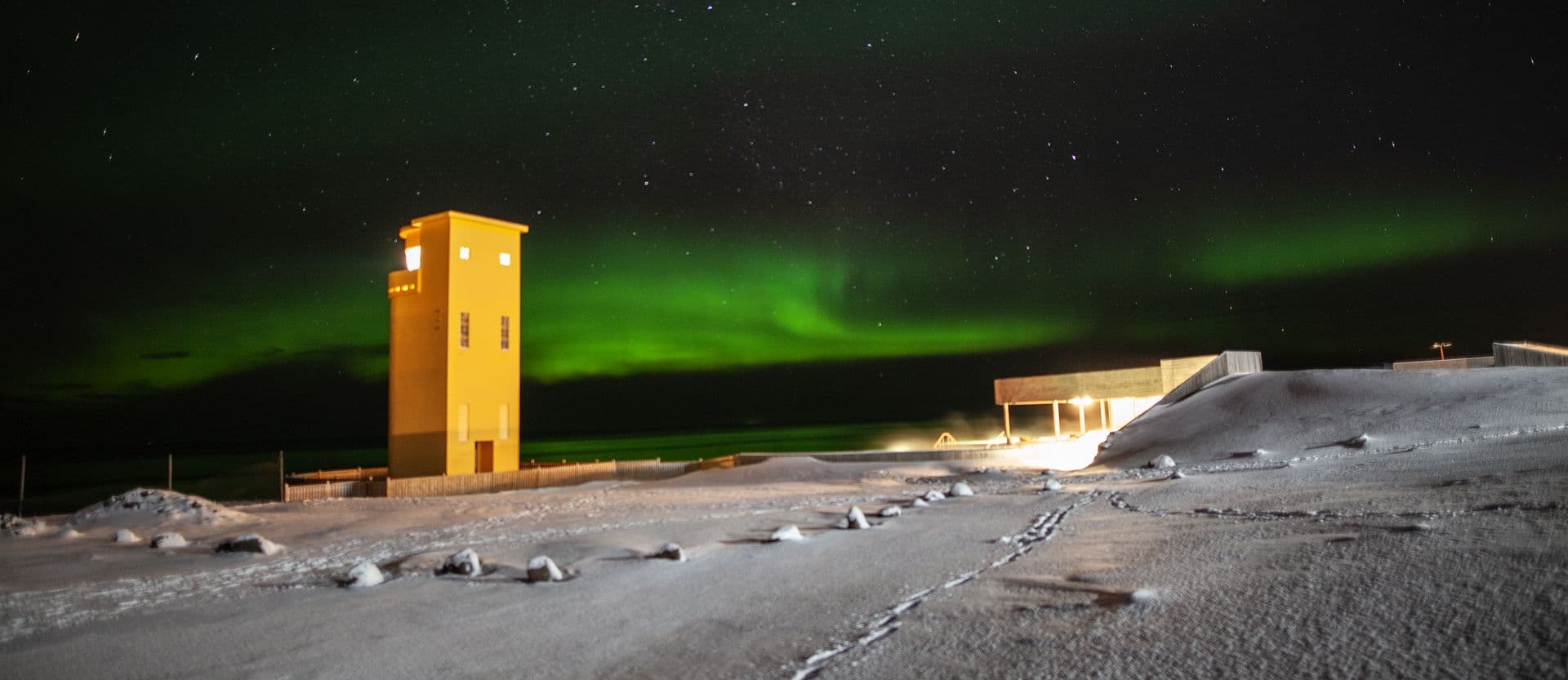 Green aurora over the yellow Húsavík lighthouse and the snowy grounds of the baths